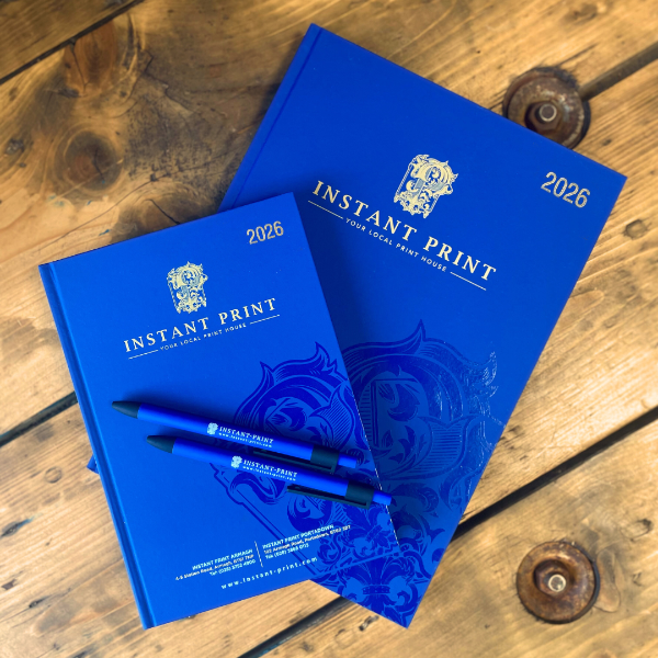 Two navy diaries with a pen on a wooden surface, featuring 'Instant Print' branding.