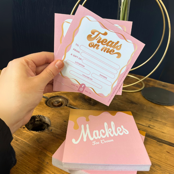 Hand holding a pink and gold 'Treats on me' gift voucher with a wooden surface in the background