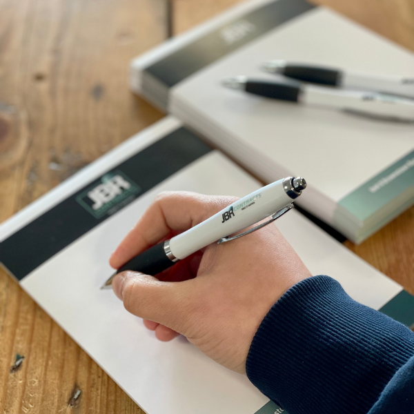 Hand holding a pen over a notepad with another pen on a wooden surface