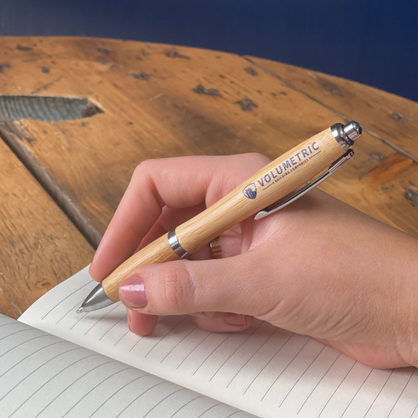 Hand holding a wooden pen with metal accents, writing in a notebook on a wooden surface.