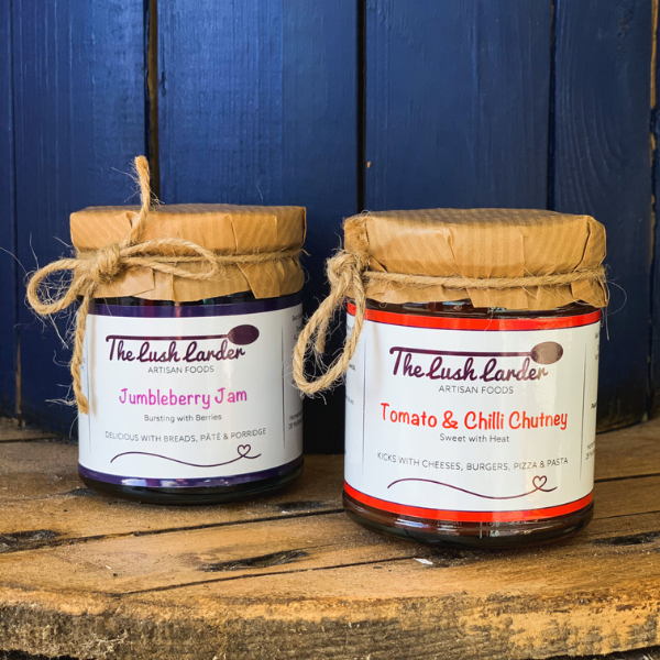 Two jars of 'The Lush Larder' jam and chutney on a wooden surface with a blue wooden background.