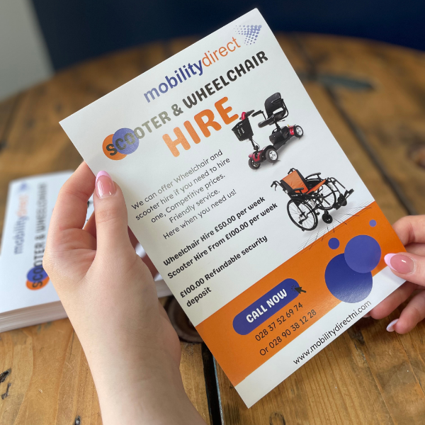 Hand holding a 'Mobility Direct' leaflet about scooter and wheelchair hire on a wooden surface.